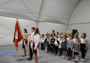Chłopiec trzyma sztandar szkoły, po jego dwóch stronach stoją dziewczynki, z tyłu grupa dzieci z chóru szkolnego