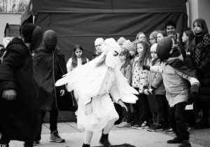 dziecko w stroju białego orła, w otoczeniu postaci ubranych w czarne stroje