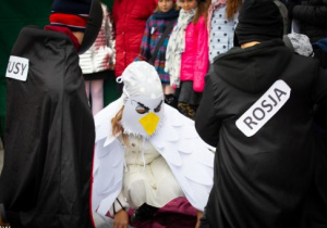 postacie przebrane w czarne stroje, symbolizujące zaborców Polski, otaczają białego orła