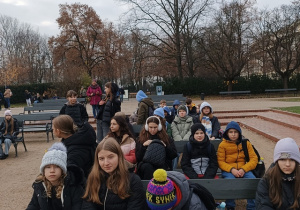 Uczniowie siedą na ławeczkach pod pomnikiem Chopina.