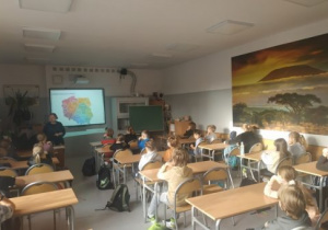 Uczniowie siedzą w ławkach szkolnych i oglądają na dużym ekranie prezentację