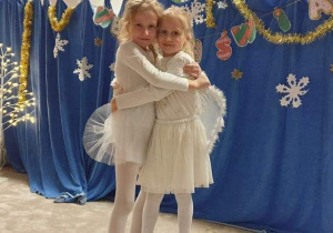 Dwie dziewczynki, grające anioły, w białych strojach ze skrzydłami, przytulone do siebie.