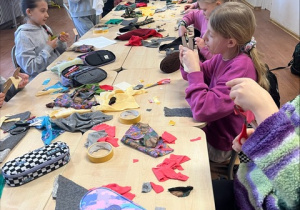 Uczniowie siedzą przy stolikach i tworzą różne stroje z otrzymanych materiałów.