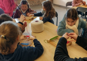 Uczniowie siedzą za stołem podczas warsztatów, tkają na małych krosnach