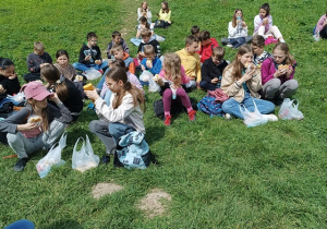 Grupa uczniów siedzi na trawie i je śniadanie.