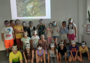 Uczniowie klasy 3b w maskach wilków, na tle ściany, na której wyświetlany jest napis: Chrońmy wilki.