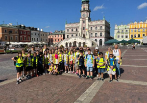 Uczniowie klasy 5a i 5b wraz z opiekunami pozują do zdjęcia na Rynku w Zamościu, w tle zabytkowy Ratusz i kamieniczki.